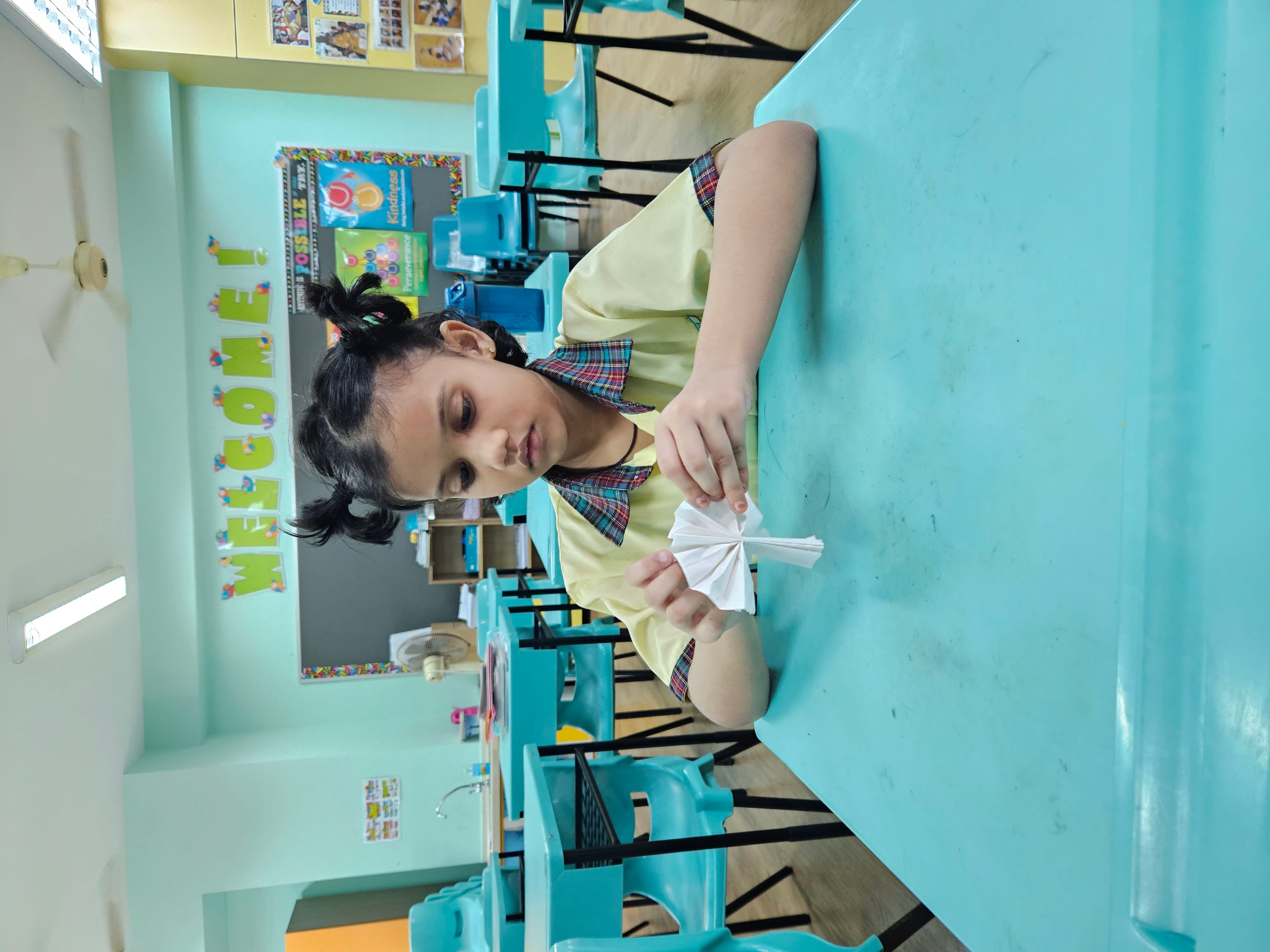 A young girl in a yellow school uniform folds a white paper craft at a teal desk in a classroom.